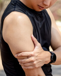 A person wearing a sleeveless black shirt and a smartwatch holds their left elbow with their right hand, appearing to experience discomfort or pain in their arm.