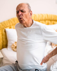 An elderly man sitting on a bed, wearing a white t-shirt, grimaces in pain while holding his lower back with one hand. The bed has yellow cushioning and white pillows in the background.