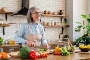 An older woman with gray hair and glasses, wearing an apron, smiles while slicing cucumber in a bright kitchen with various fresh vegetables and plants on the counter.