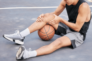 A basketball player sits on an outdoor court, holding his knee in pain with a basketball under his leg, suggesting he may have been injured during a game.