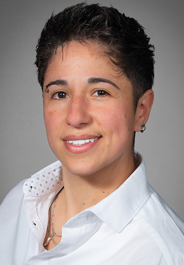 Person with short dark hair and a light complexion, smiling and wearing a white collared shirt, small hoop earring, and layered necklaces, posing against a neutral gray background.