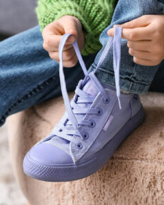 Child in blue jeans and a green sweater tying the shoelaces of a light purple sneaker, sitting on a beige textured surface.